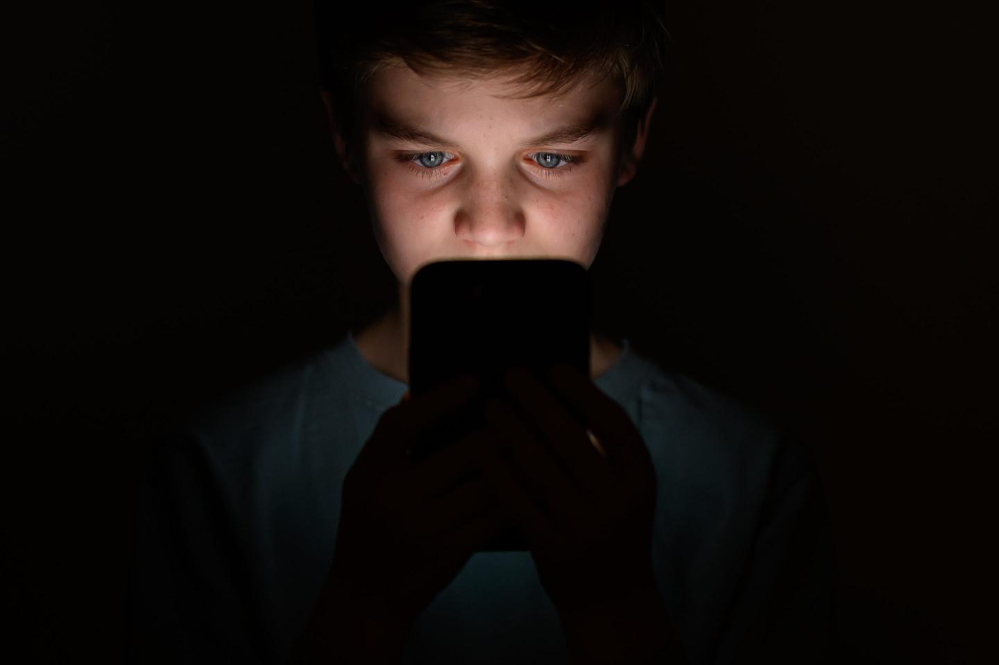 A boy stares at a smartphone against a black background