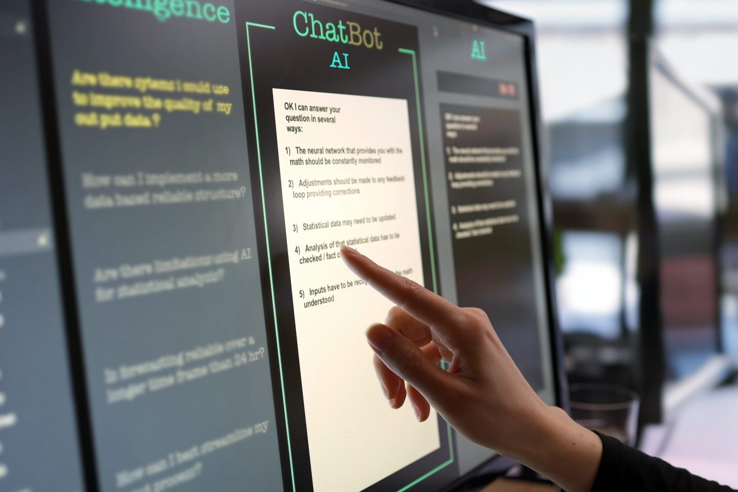 Close-up stock photograph showing a touchscreen monitor being used in an open plan office. A woman’s hand is asking an AI chatbot pre-typed questions & the Artificial Intelligence website is answering.