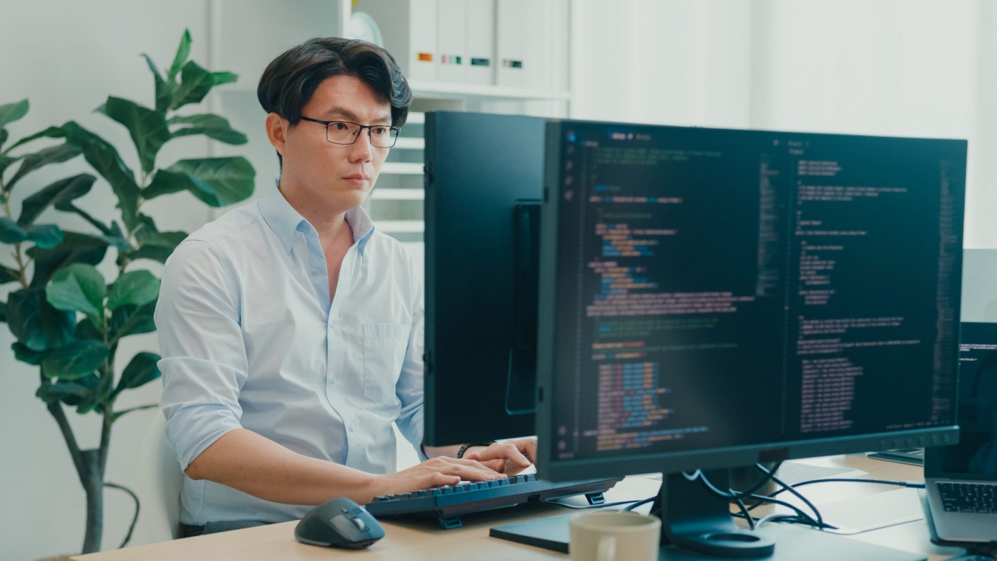 Young Asian man software developers using computer to write code sitting at desk with multiple screens work at office. Programmer development.