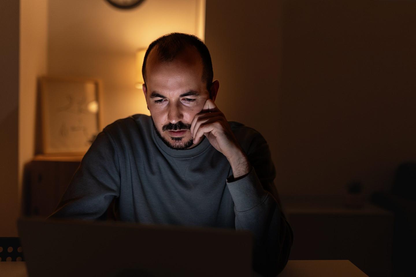 Young man looking worriedly at his laptop screen while working from home at night. People and home office concept.