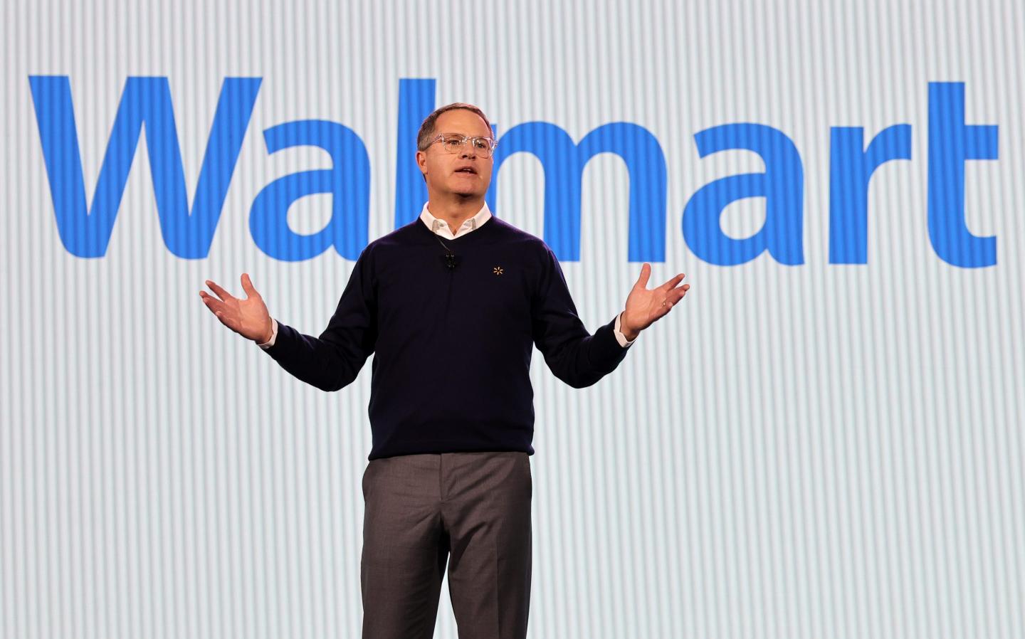 Walmart CEO Doug McMillon stands onstage with his hands spread apart, with a Walmart image displayed on a screen behind him.