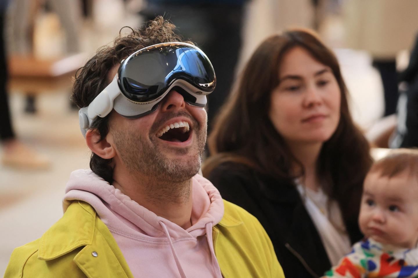 A man in a pink hoodie and yellow jacket is wearing the Apple Vision Pro, looking up, and smiling in awe.