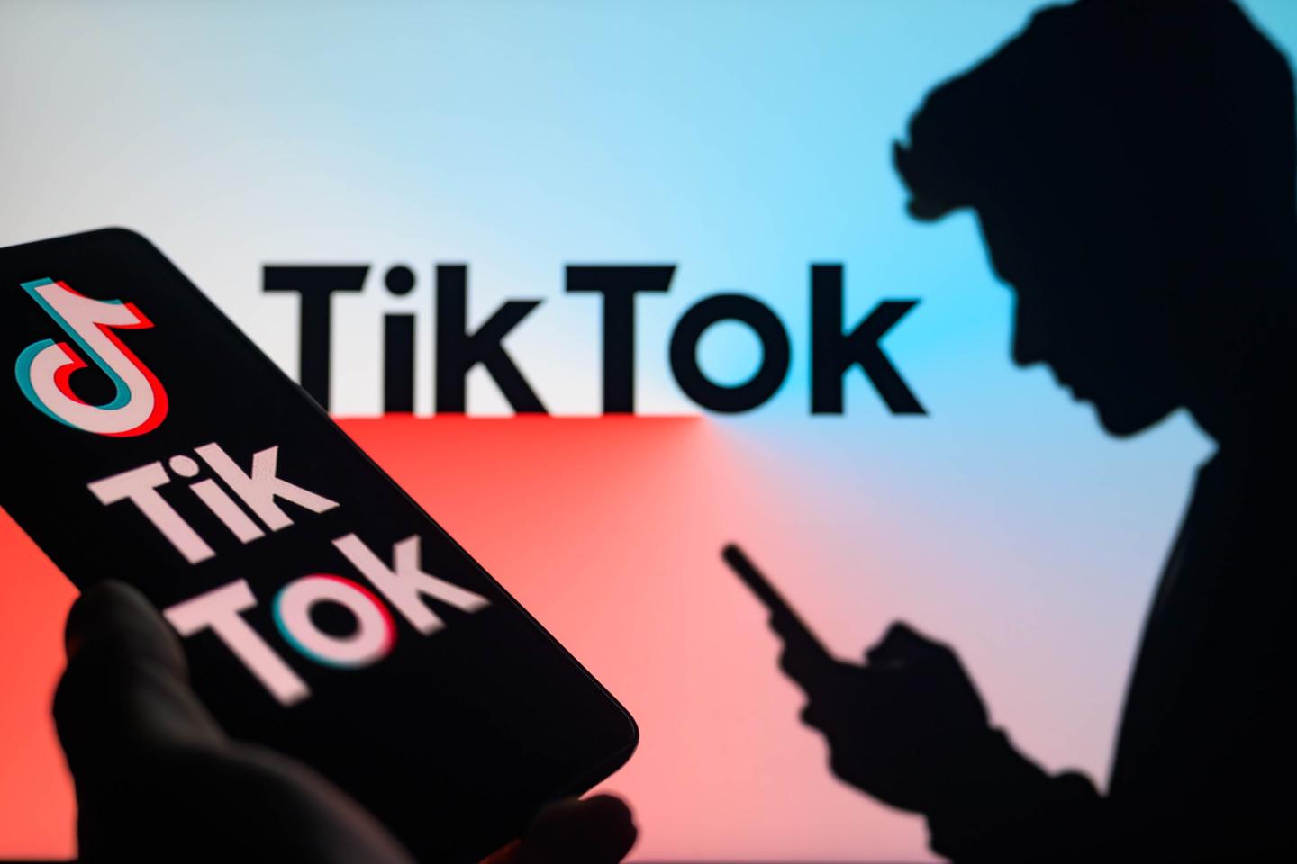 The TikTok logo is being displayed on a smartphone with TikTok visible in the background in this photo illustration