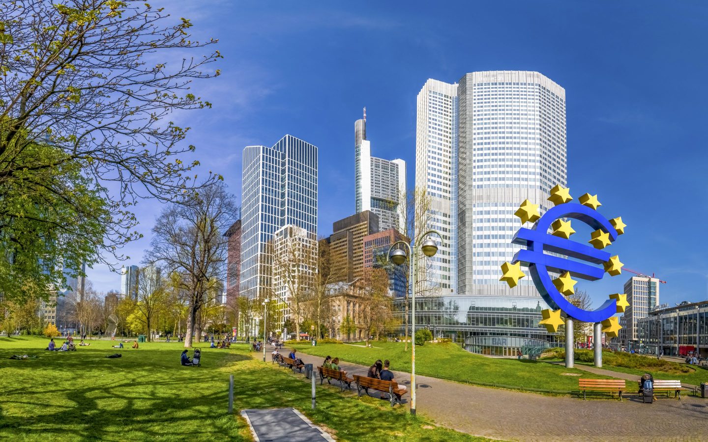 people sitting on benches at a park with a big blue European Central Bank logo
