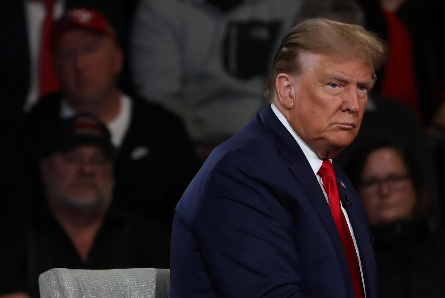 Trump is sitting sideways and has a serious expression. He’s wearing a dark suit, white shirt and red tie.