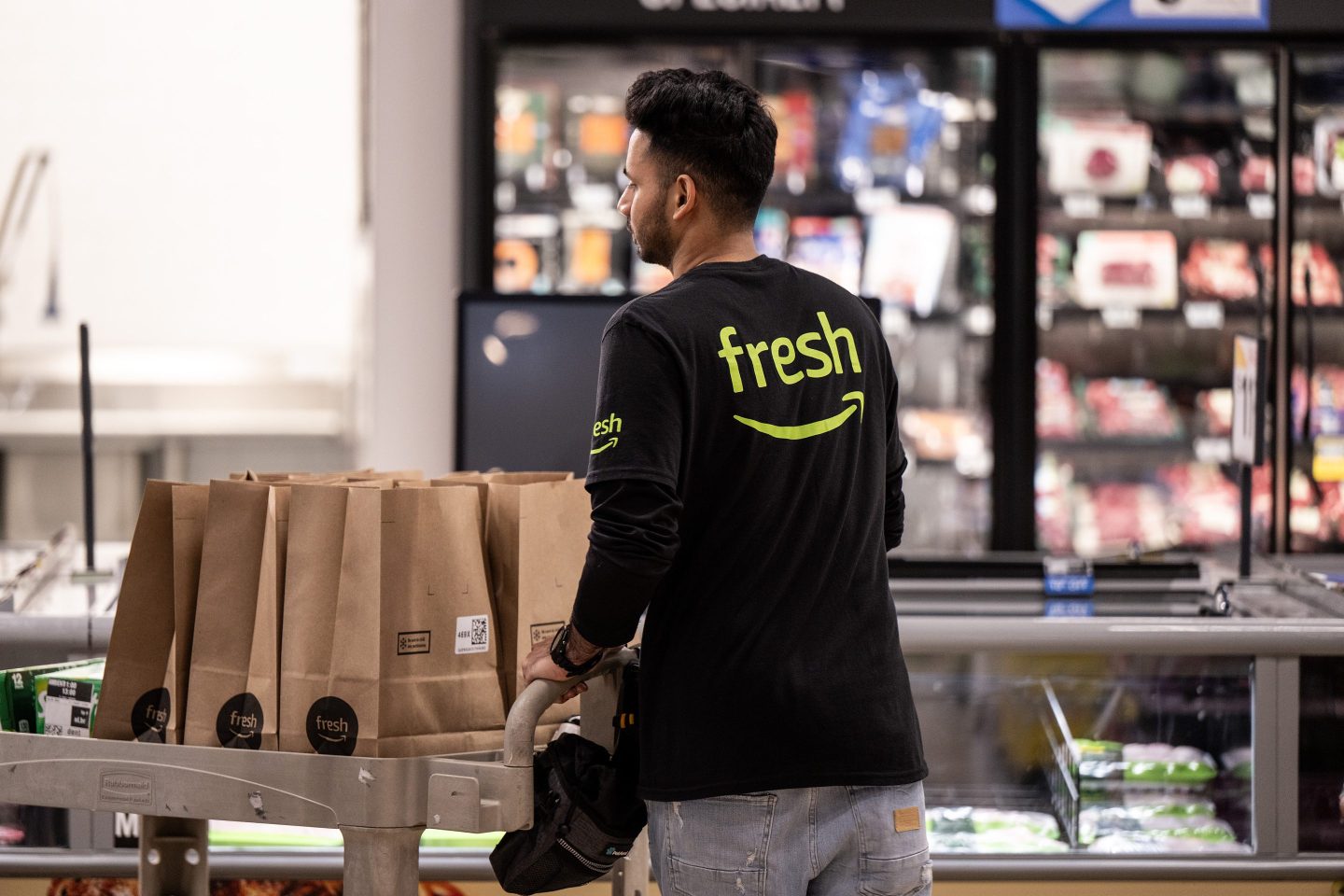 An Amazon worker pushes a cart full of grocery orders in paper bags across an Amazon Fresh store.