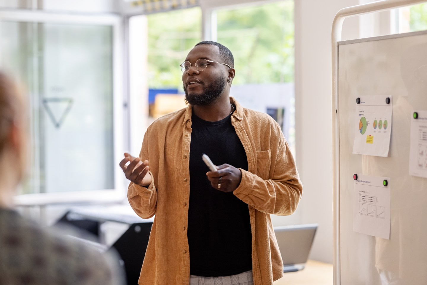 African man giving a presentation to the team at coworking office, standing by a whiteboard with statistical reports explaining new strategies during meeting.