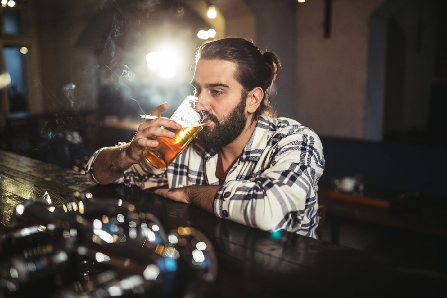 A bearded man is sitting at a bar drinking beer with a lit cigarette in his hand.