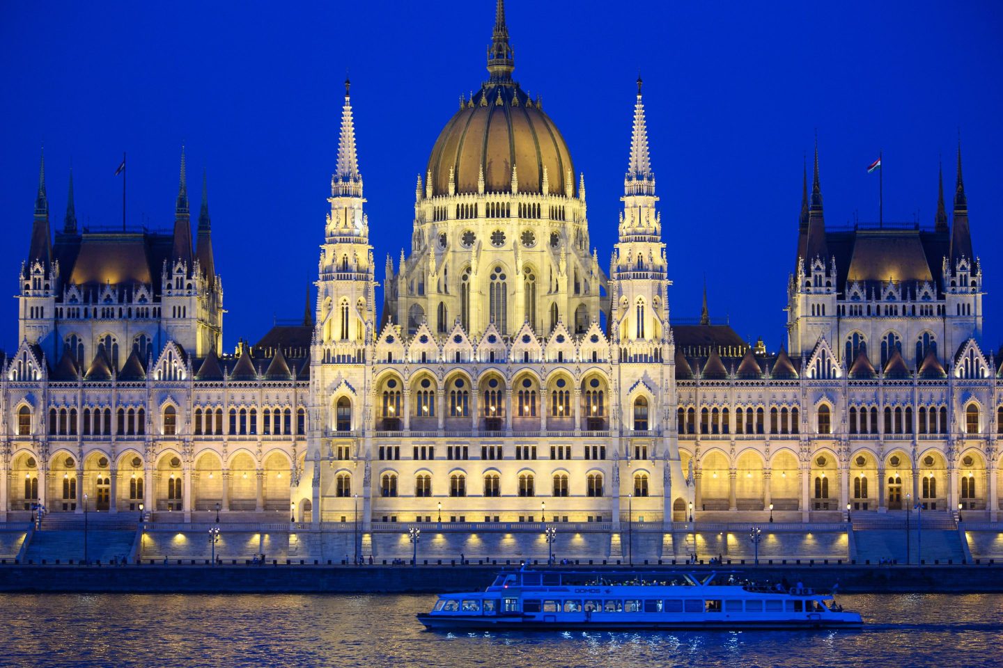 An excursion boat sails along the Danube in the evening past the illuminated parliament building.
