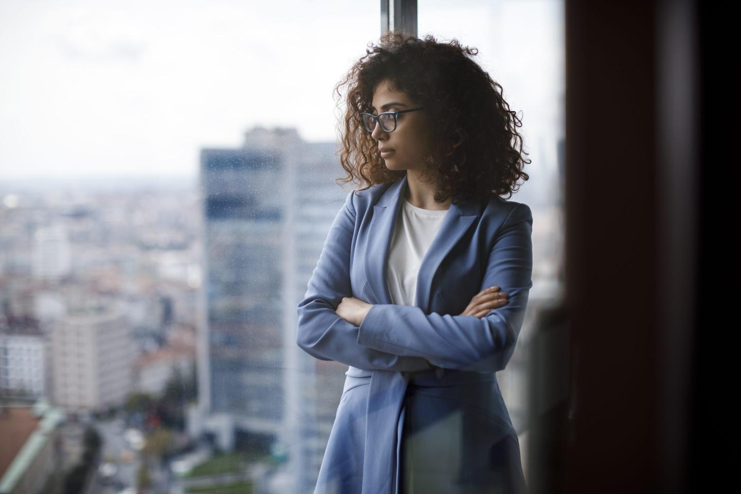 A young woman dressed in business attire gazes glumly through a high-rise office window.