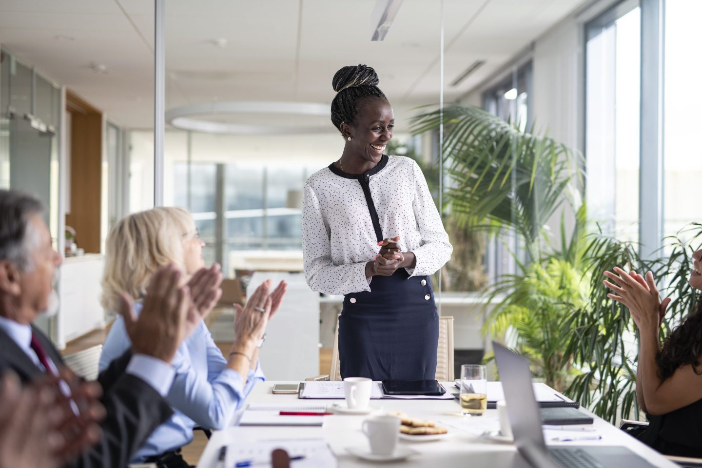 A young Black businesswoman stands in front of her colleagues at the head of a board room meeting table. Her colleagues applaud her.