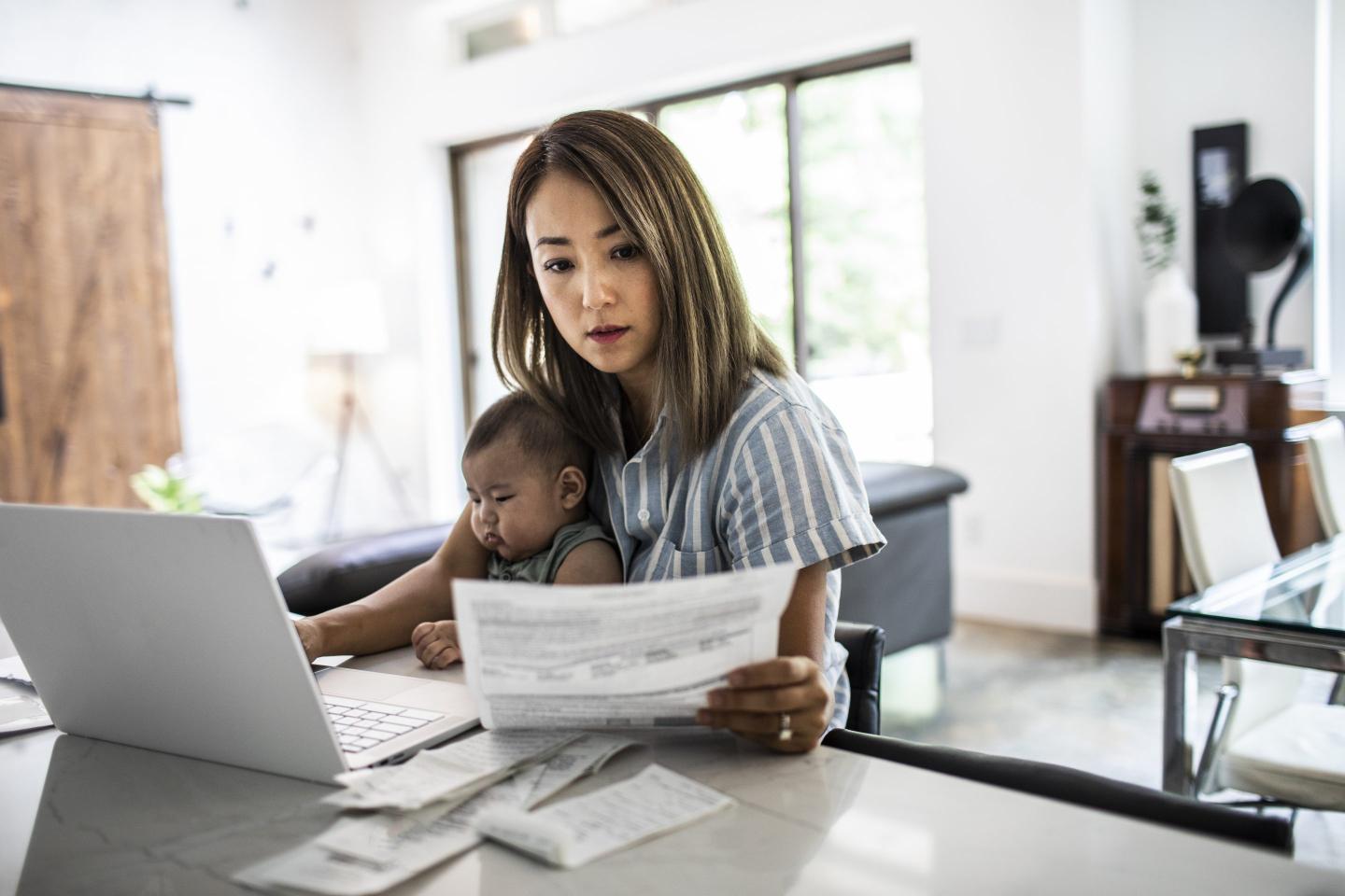 Woman at laptop holding baby and looking at documents