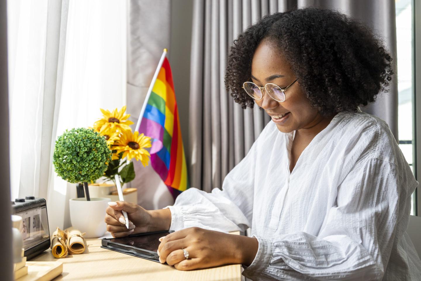 A young woman works on a tablet at a desk. There is a pride flag behind her.