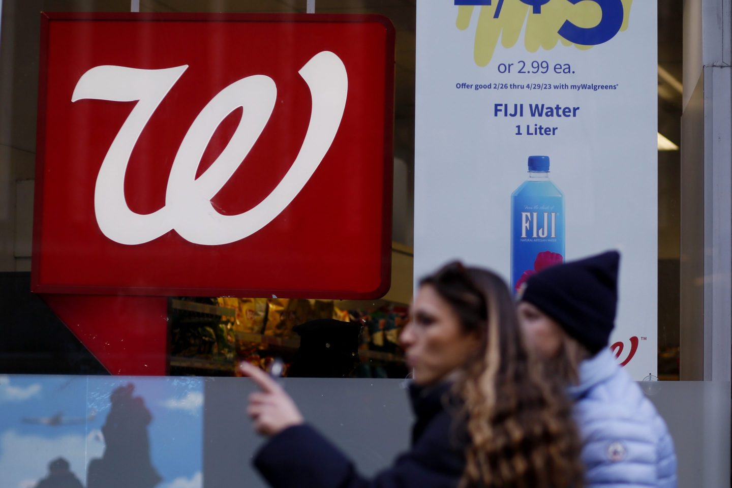 An out-of-focus person is point to their left and is standing in front of a window with the red and white Walgreens 'W' on it.