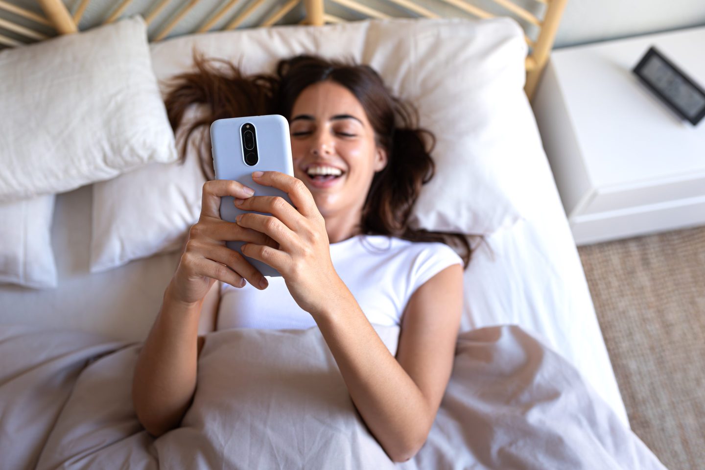 Happy young woman using mobile phone in bed.