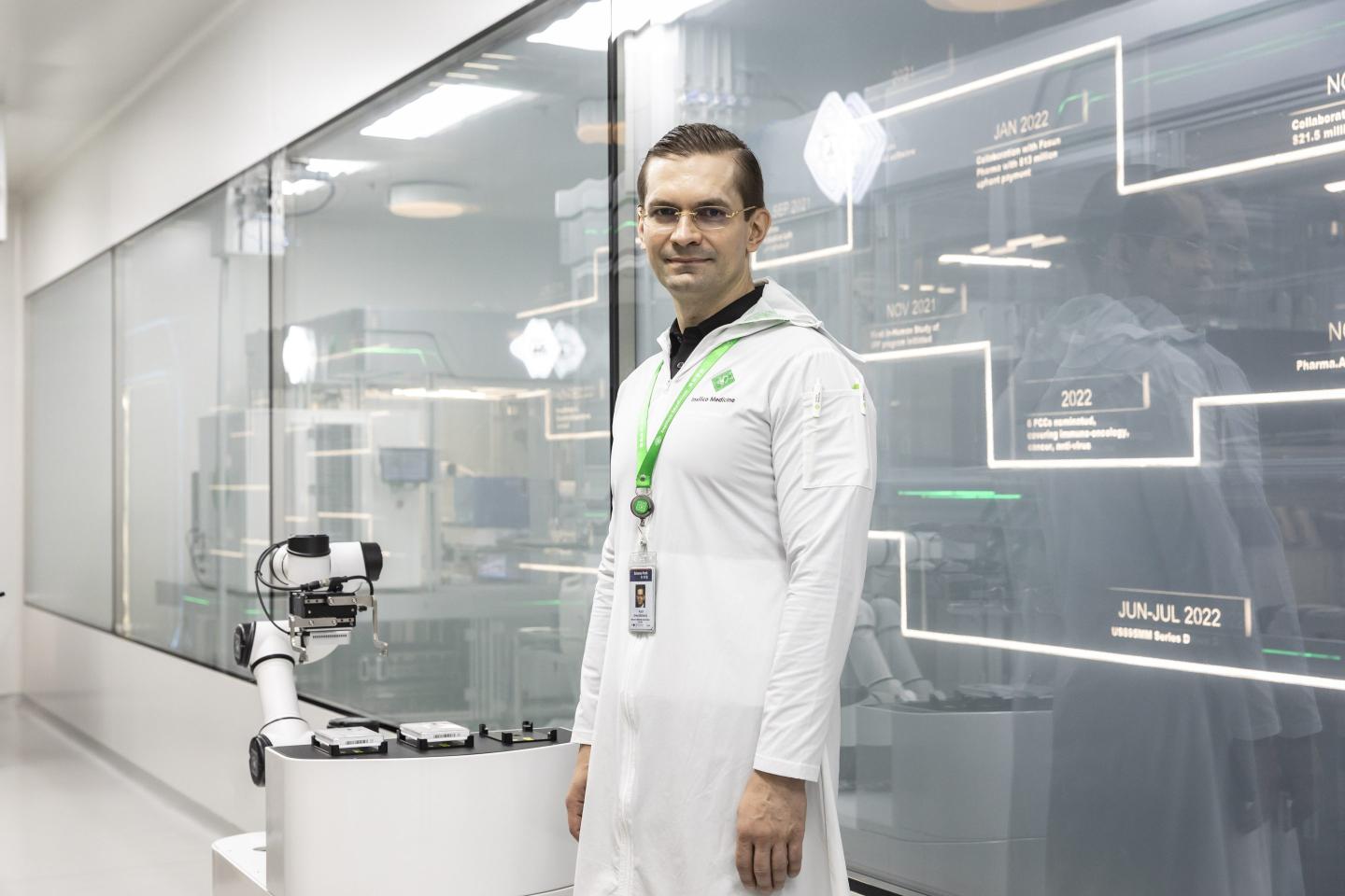 A man with glasses and a white lab coat stands in front of a glass wall in a medical research facility.