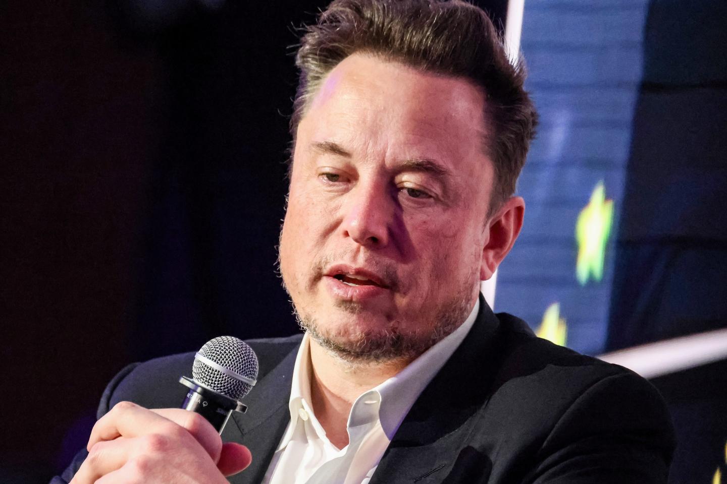 Elon Musk is wearing a suit a holds a microphone as he is speaking.