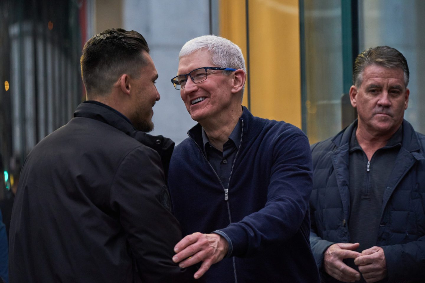 A white-haired older man wearing glasses, smiles and hugs another man.