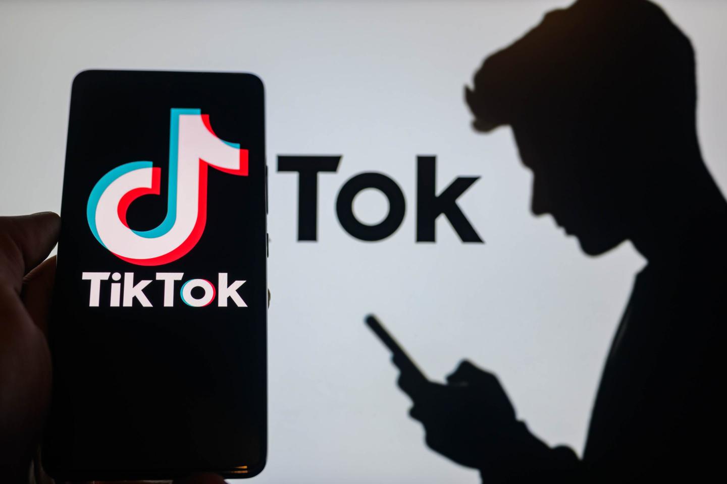 The TikTok logo is being displayed on a smartphone with TikTok visible in the background