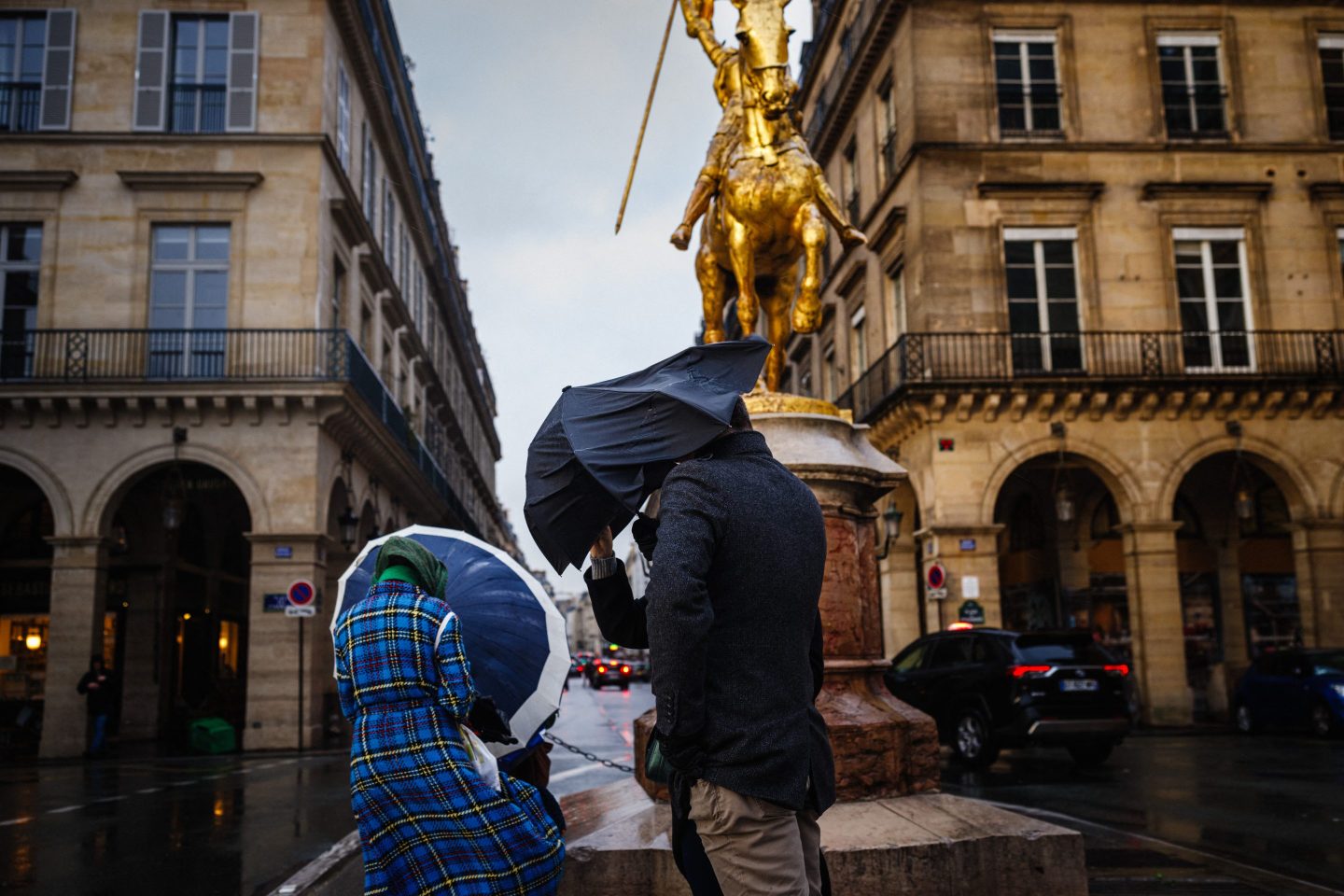 Pedestrians shelter from the rain and the wind under umbrellas, as they walk by the sculpture depicting Joan of Arc in France.