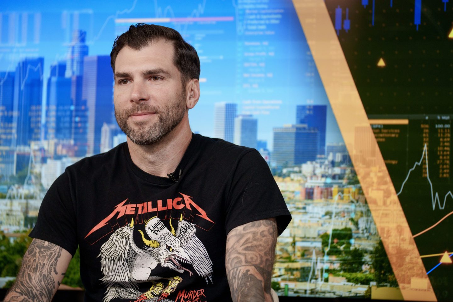 Mike Cessario is wearing a black Metallica T-shirt and is sitting in front of a background overlooking a skyline.