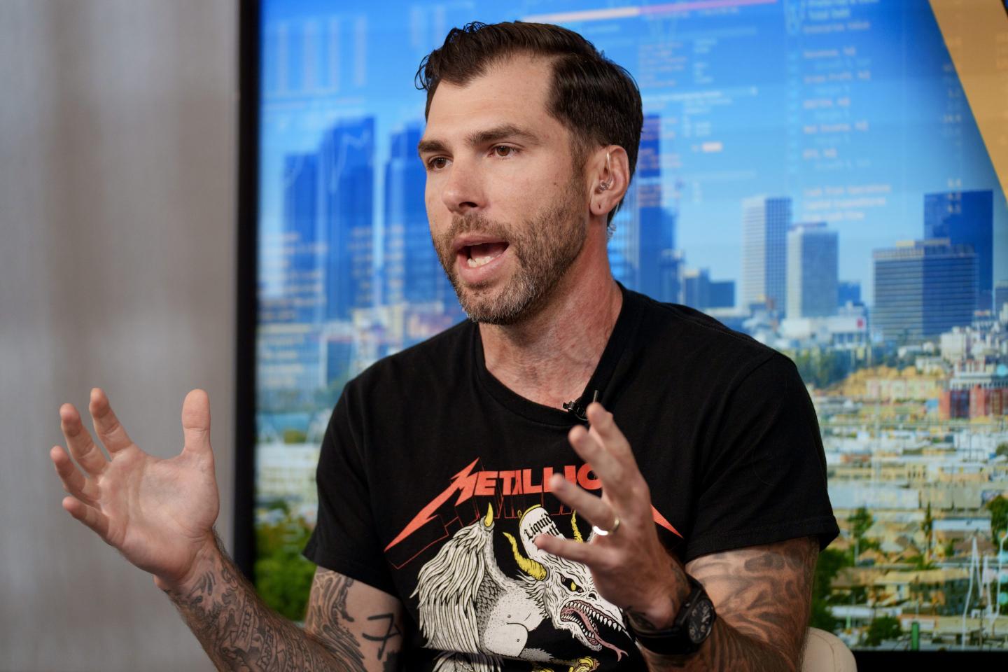 Mike Cessario is wearing a black Metallica T-shirt while speaking in front of a window looking out over skyscrapers.
