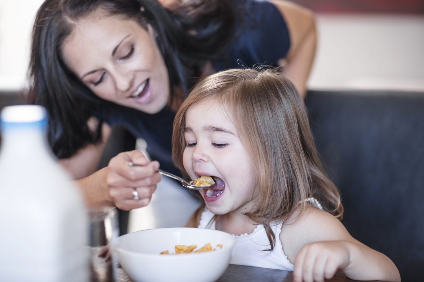 A woman is leaning over the chair a girl is sitting at and is spooning cereal into the girl's mouth.