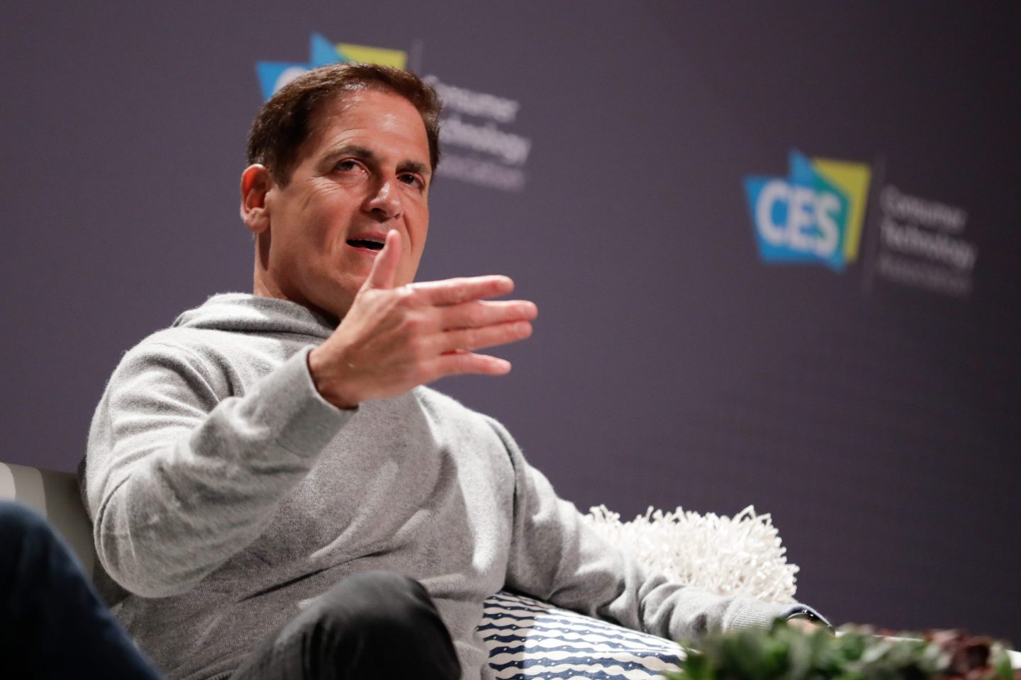 Mark Cuban is seated wearing a light gray hoodie with his right hand extended.