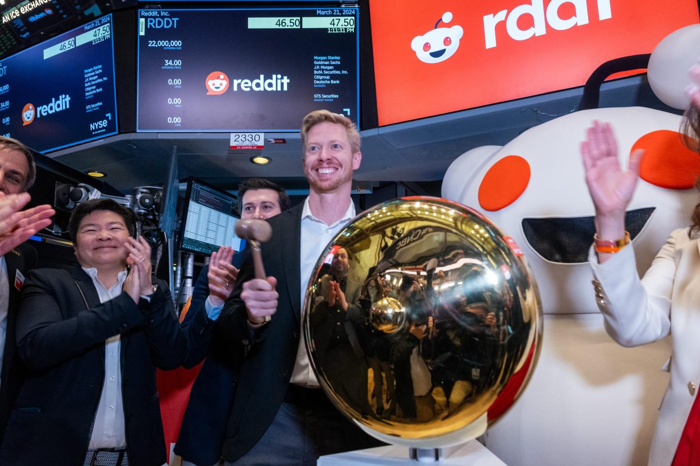 Reddit CEO Steve Huffman is wearing a sports and a button down shirt. He is holding a hammer to ring the gold bell at the center of the picture. Also in the background is Reddit's mascot, Snoo.