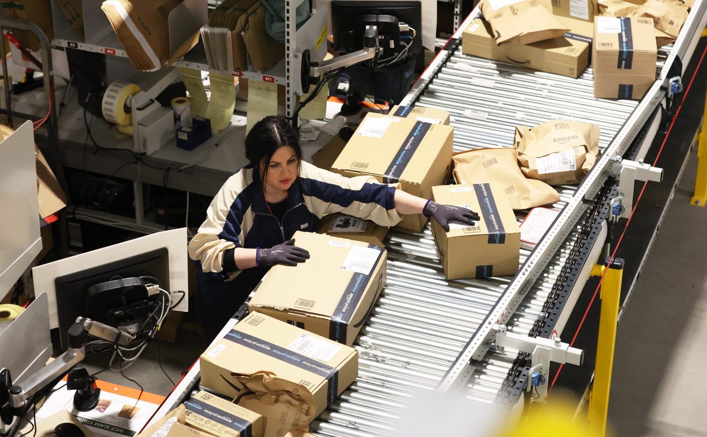 A female Amazon worker sorts customer orders in boxes that are traveling along a conveyor belt.
