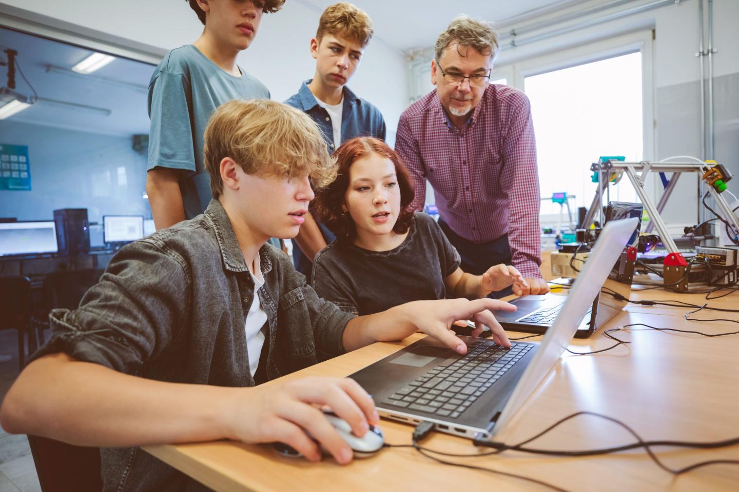 Group of students learn how to code around a laptop as a teach helps.