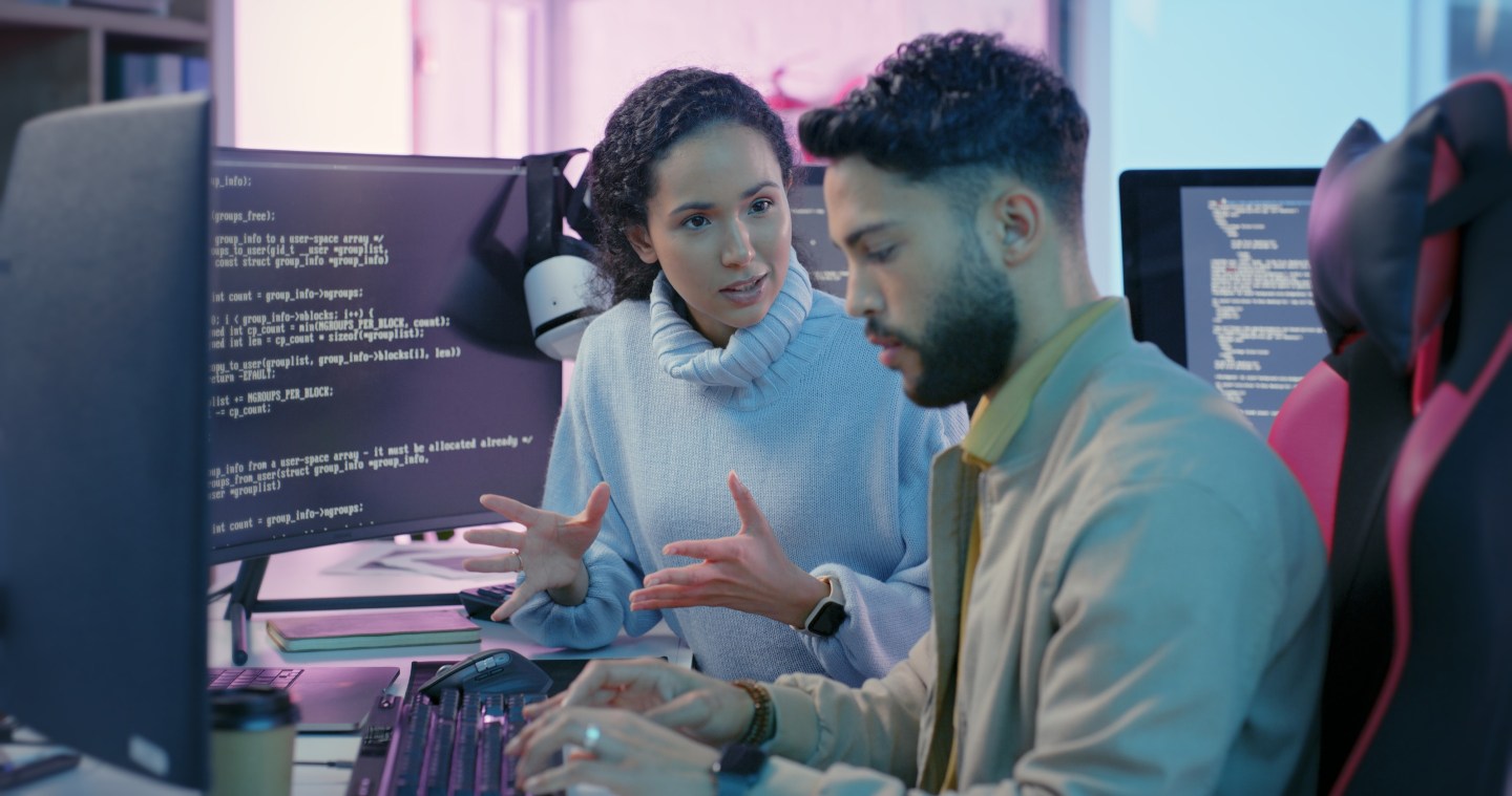 Developer explains a concept to another developer, both sitting in front of computer screens