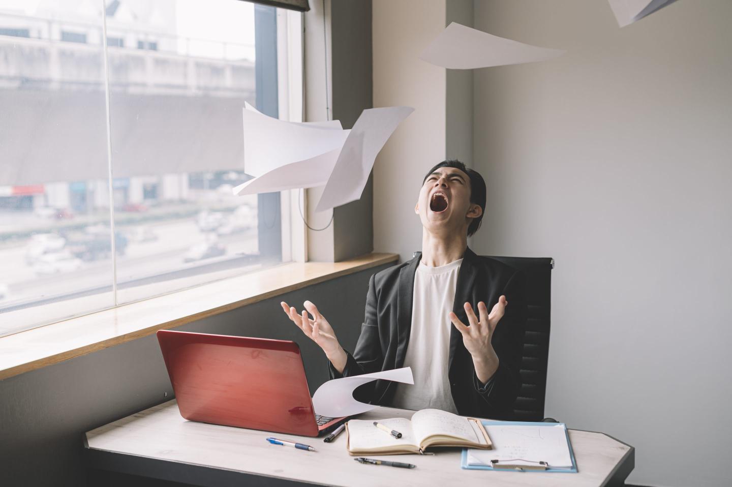 A young man sitting at his computer throws a stack of paper in the air and yells