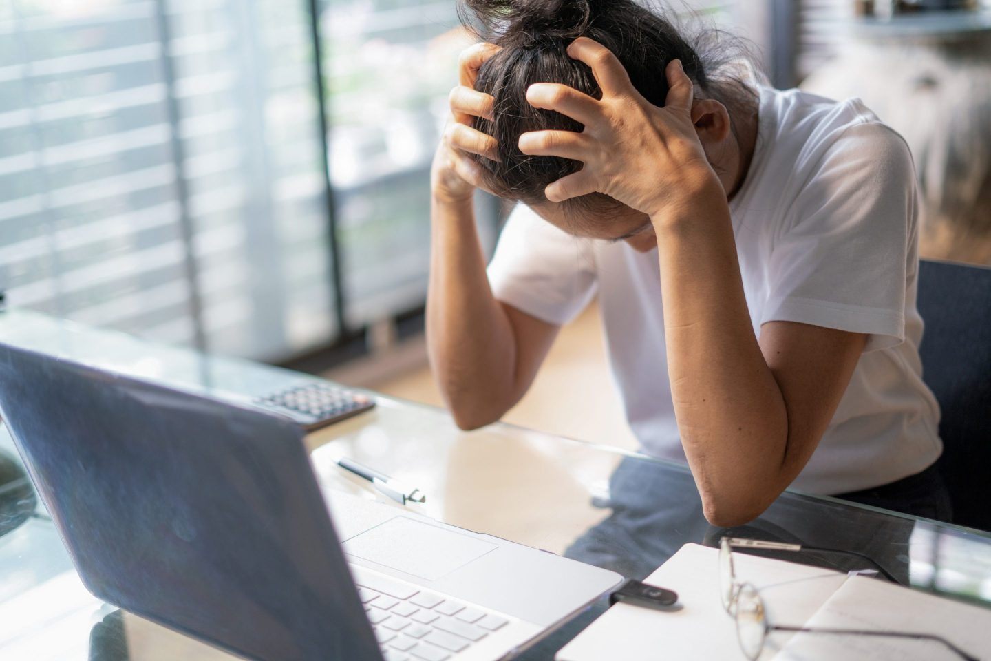 A young woman looks stressed as she cradles her head in her hands while sitting at a desk with a computer in front of her.