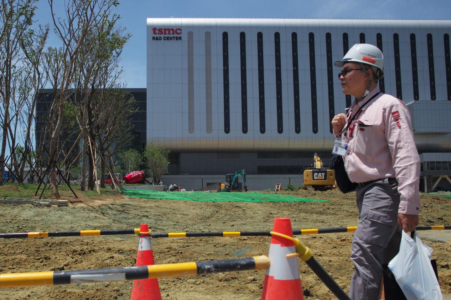 A man wearing a hardhat walks across the courtyard in front of a TSMC building.