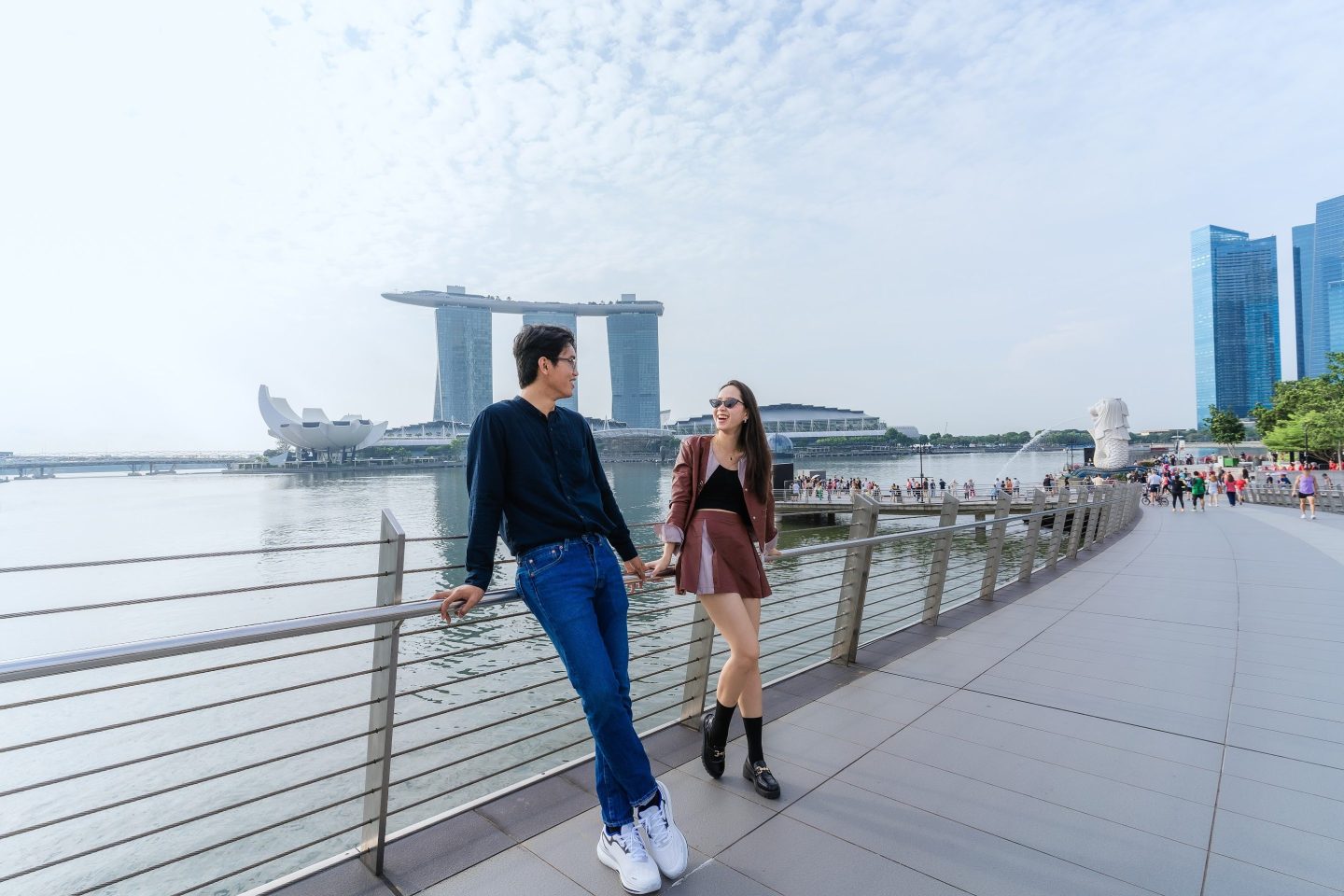 Tourist couple walking along Marina Bay with merlion park and buildings in the background.