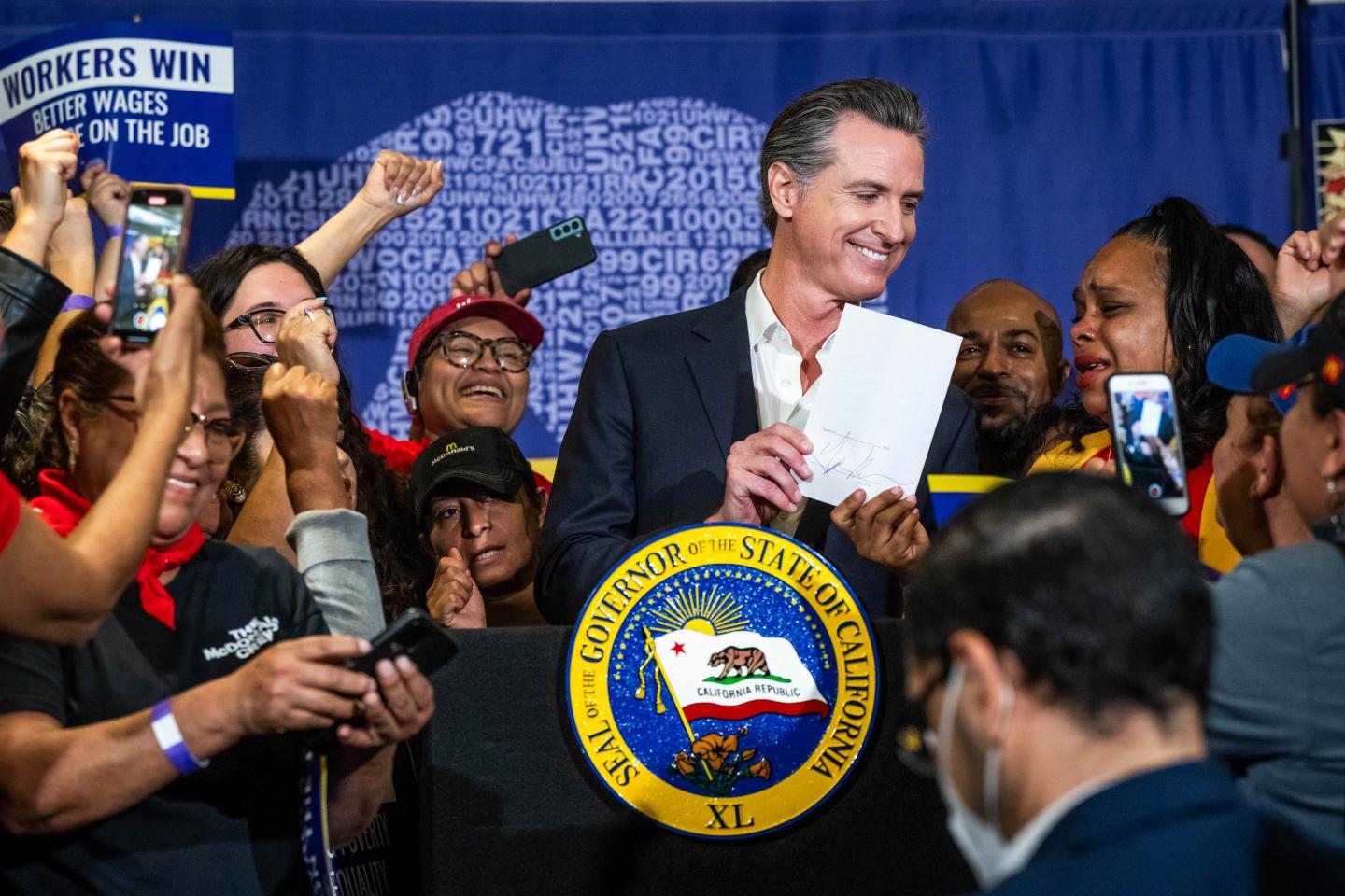 Gavin Newsom smilies with a piece of paper behind a podium. He is surrounded by celebrating workers and supporters.