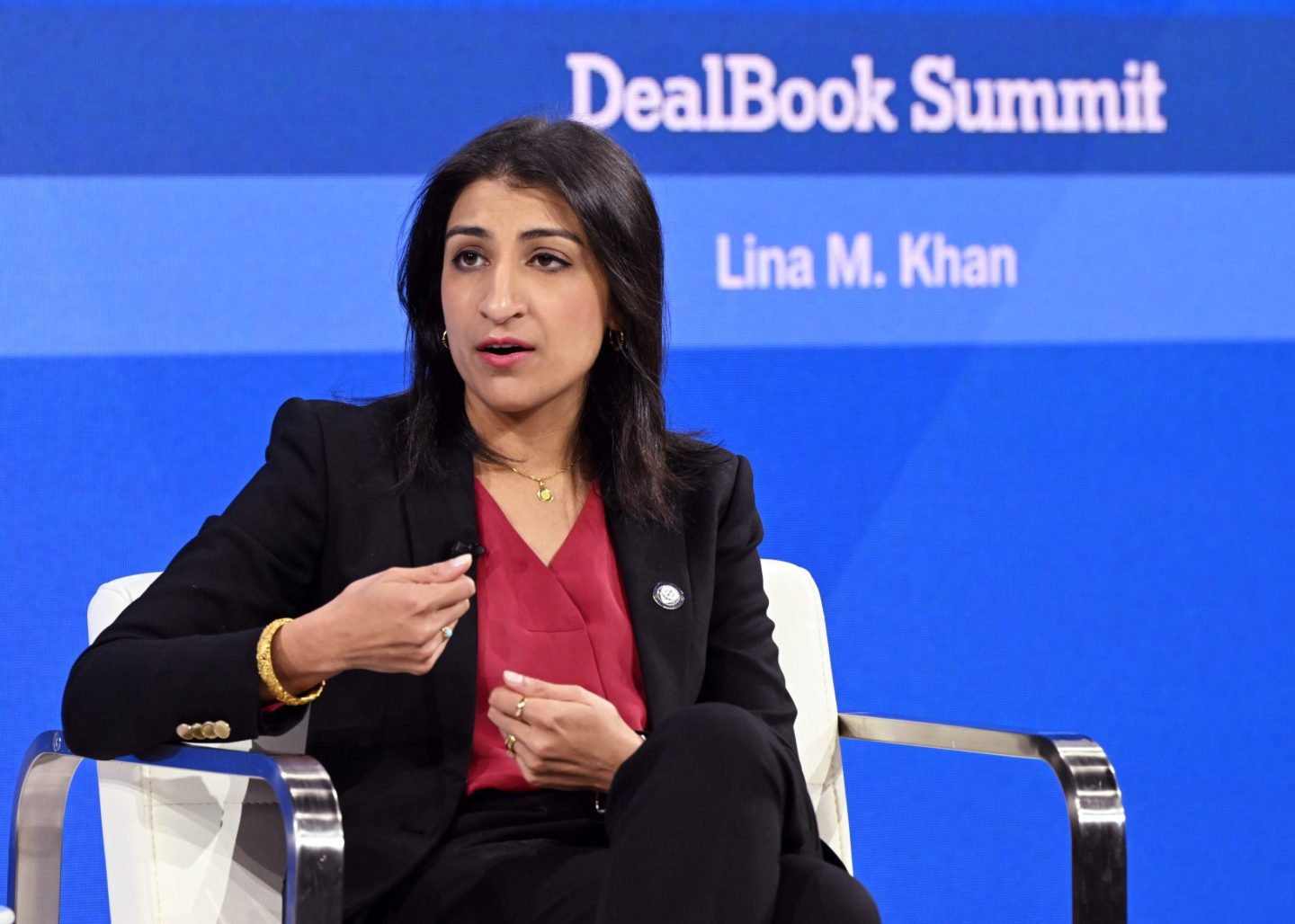 FTC Chair Lina Khan is sitting in a white chain in front of a blue background, gesturing as she speaks.