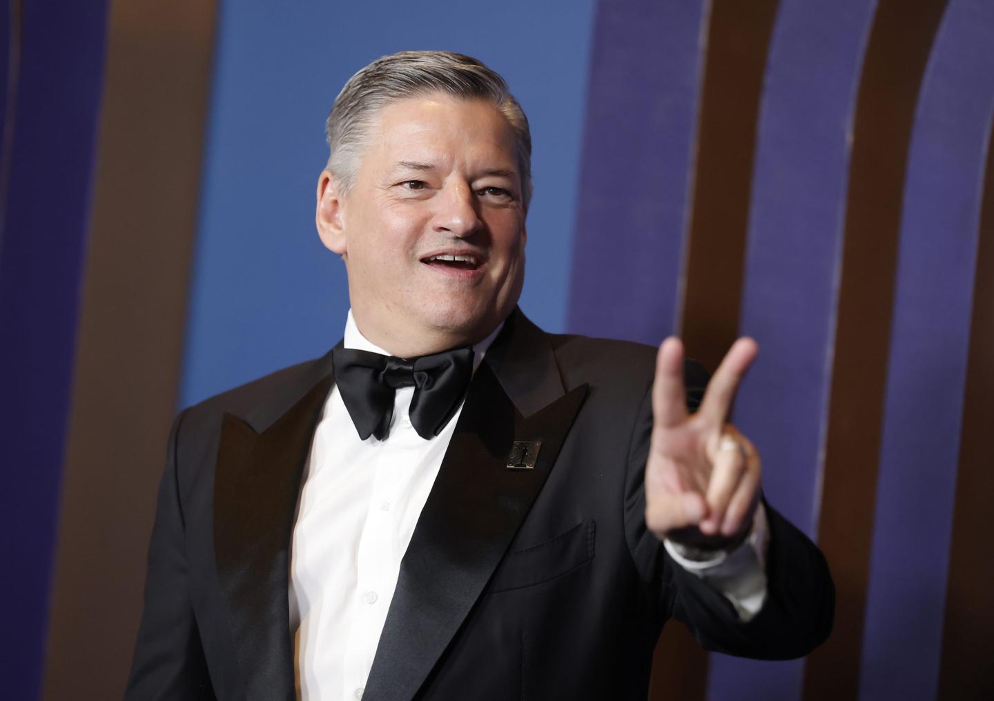 Ted Sarandos flashes a peace sign while wearing a suit.