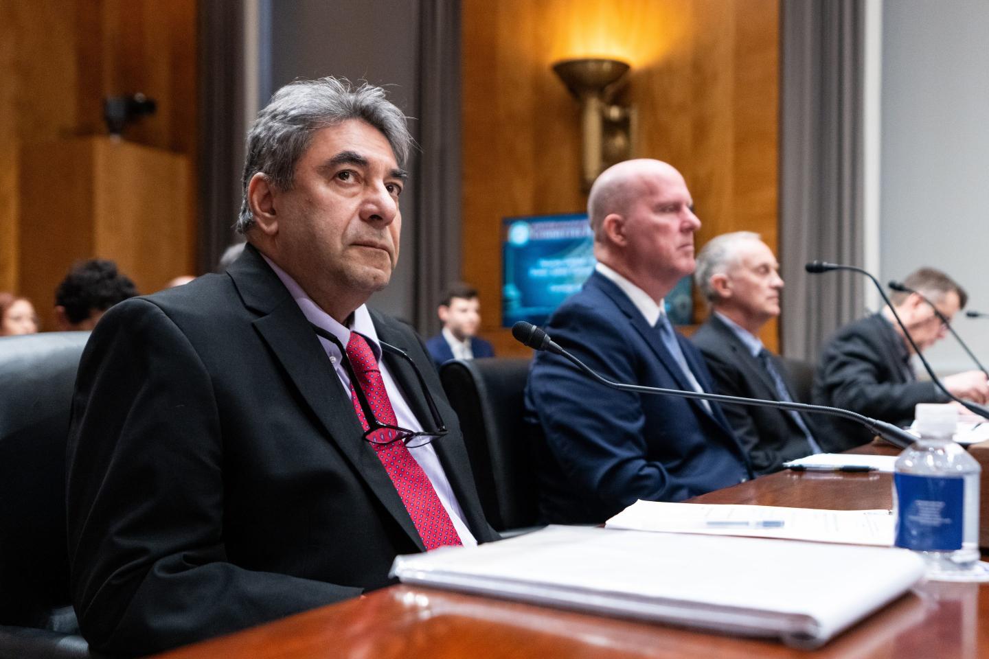 Three men in suits sit at a table with papers sprawled in front of them.