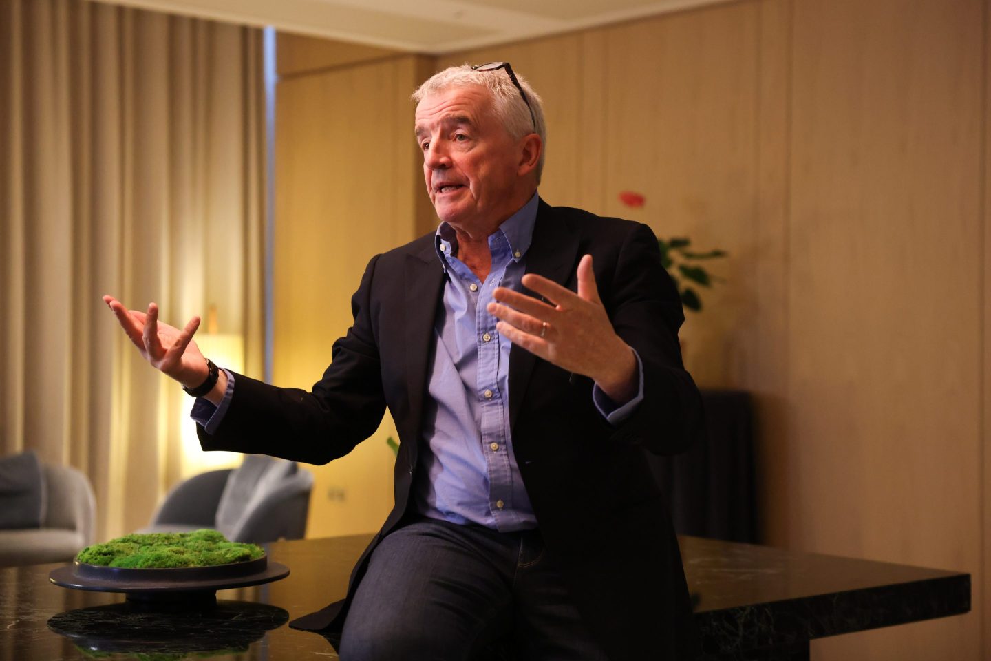 Ryanair CEO Michael O'Leary is wearing a suit and leaning on a table, gesturing as he speaks.