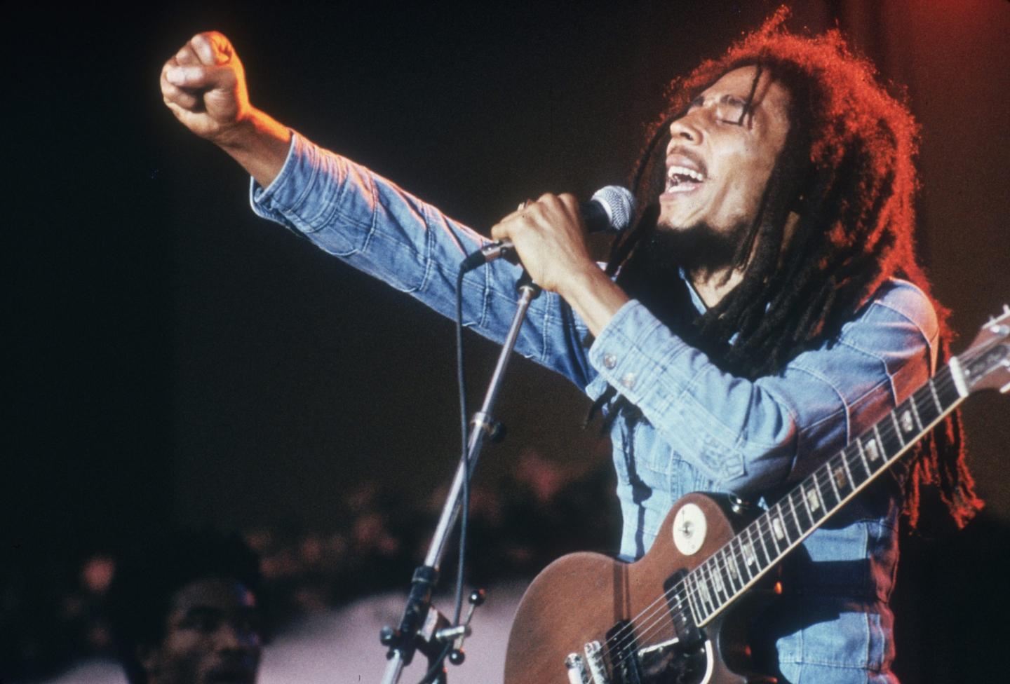 Jamaican Reggae musician, songwriter, and singer Bob Marley performs on stage, in a concert at Grona Lund, Stockholm, Sweden. He extends his fist as he sings into the microphone, with an electric guitar.