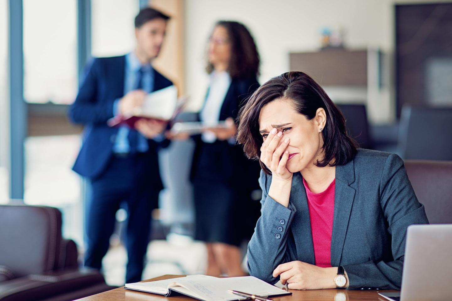 A businesswoman sitting at a desk in an office cries while her colleagues talk in the background.