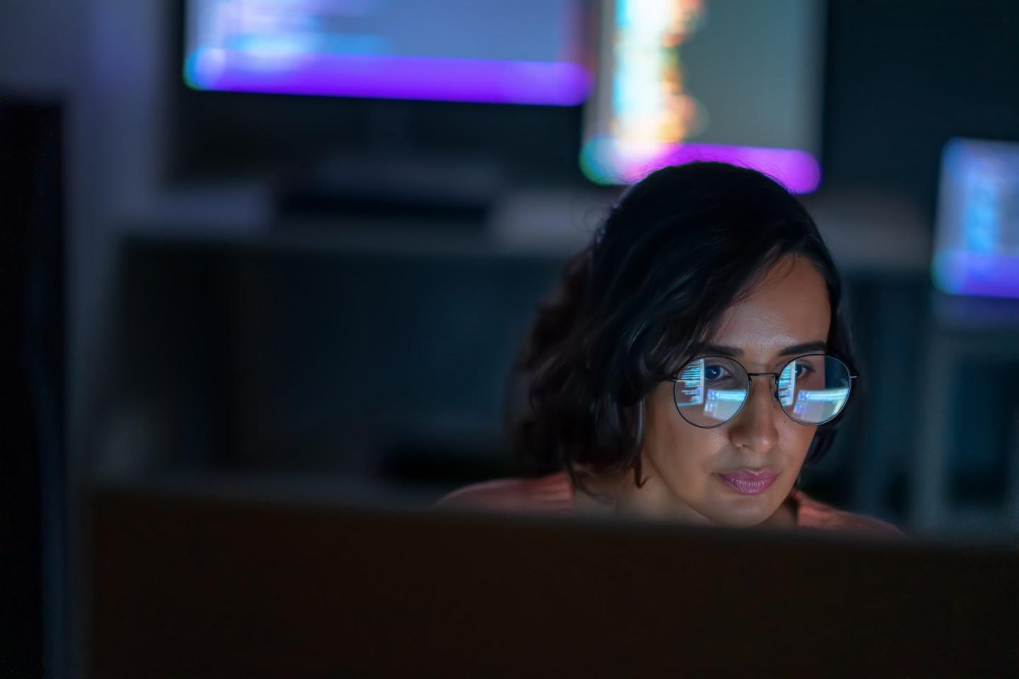 Woman in a dark room looks at screen, which is reflected in her glasses