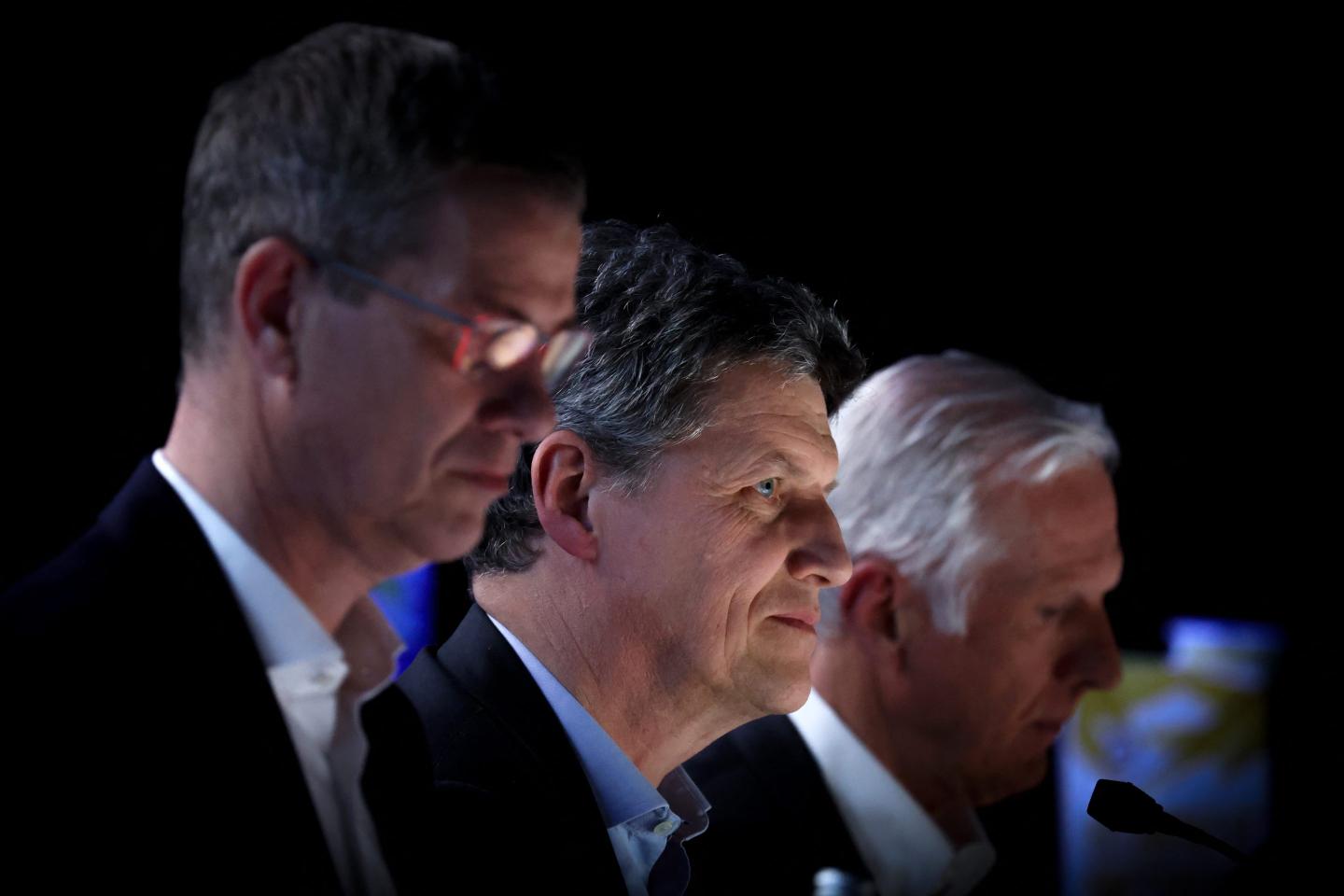Three seated executives listen to a speech