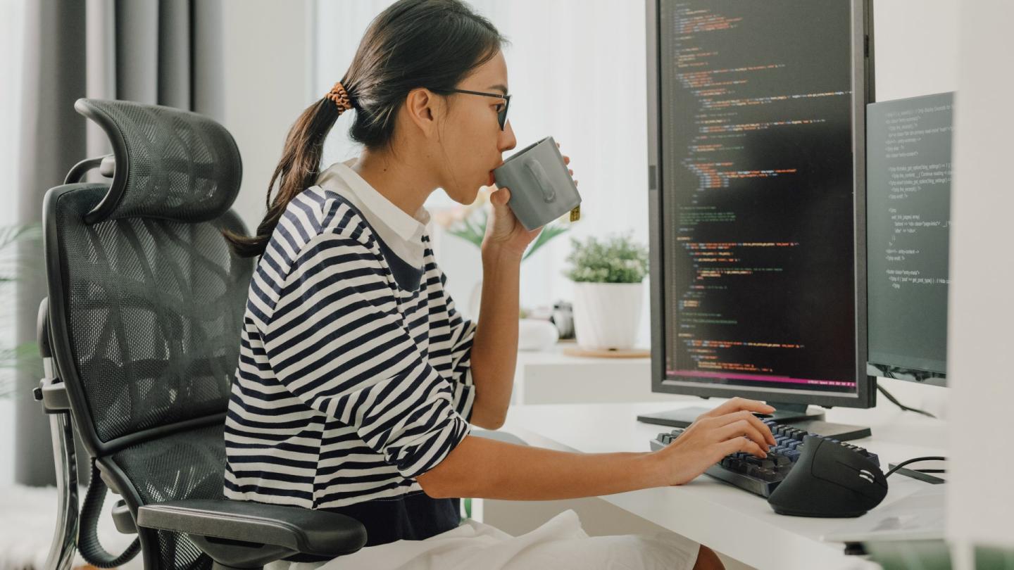 Young Asian woman is a software developer, drinking coffee and using computer to write code, sitting at desk with multiple screens work remotely at home. Striped shirt.