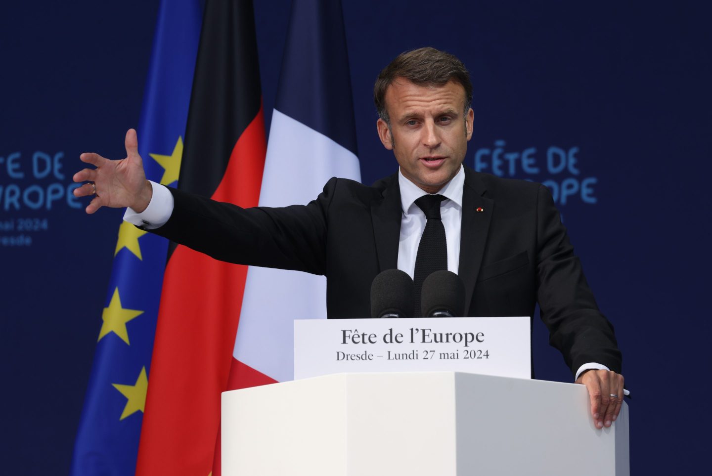 Emmanuel Macron gestures with his arm out wide during a speech at a podium with French, EU and German flags in background
