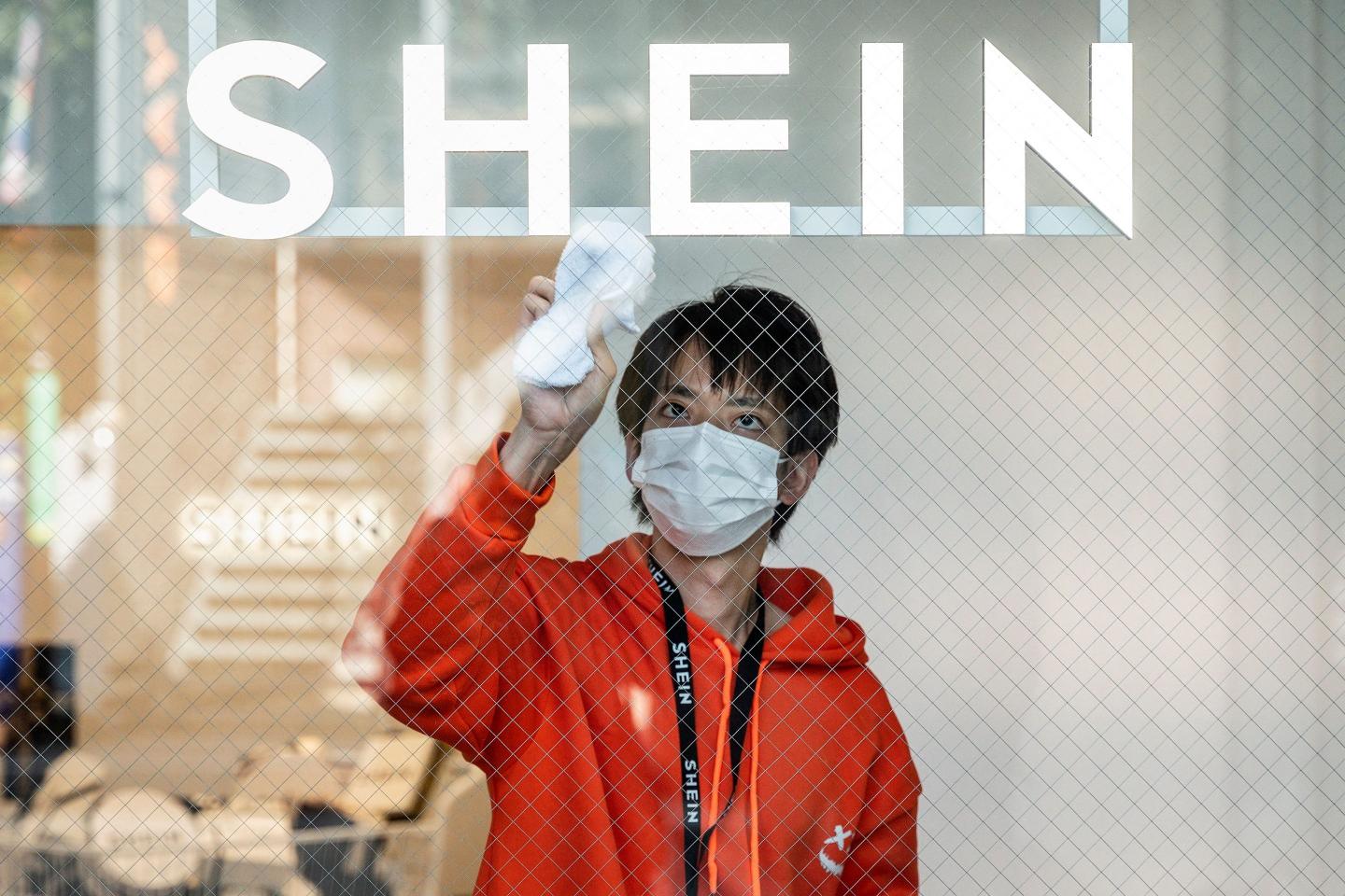 A man in a white mask and orange hoodie cleans a window with a big SHEIN sign above it.