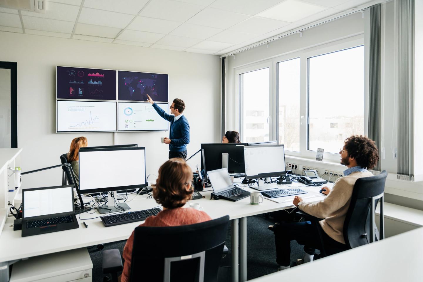 A business office with a number of employees working at computers while listening to a colleague talking and reviewing some data on wall mounted monitors.