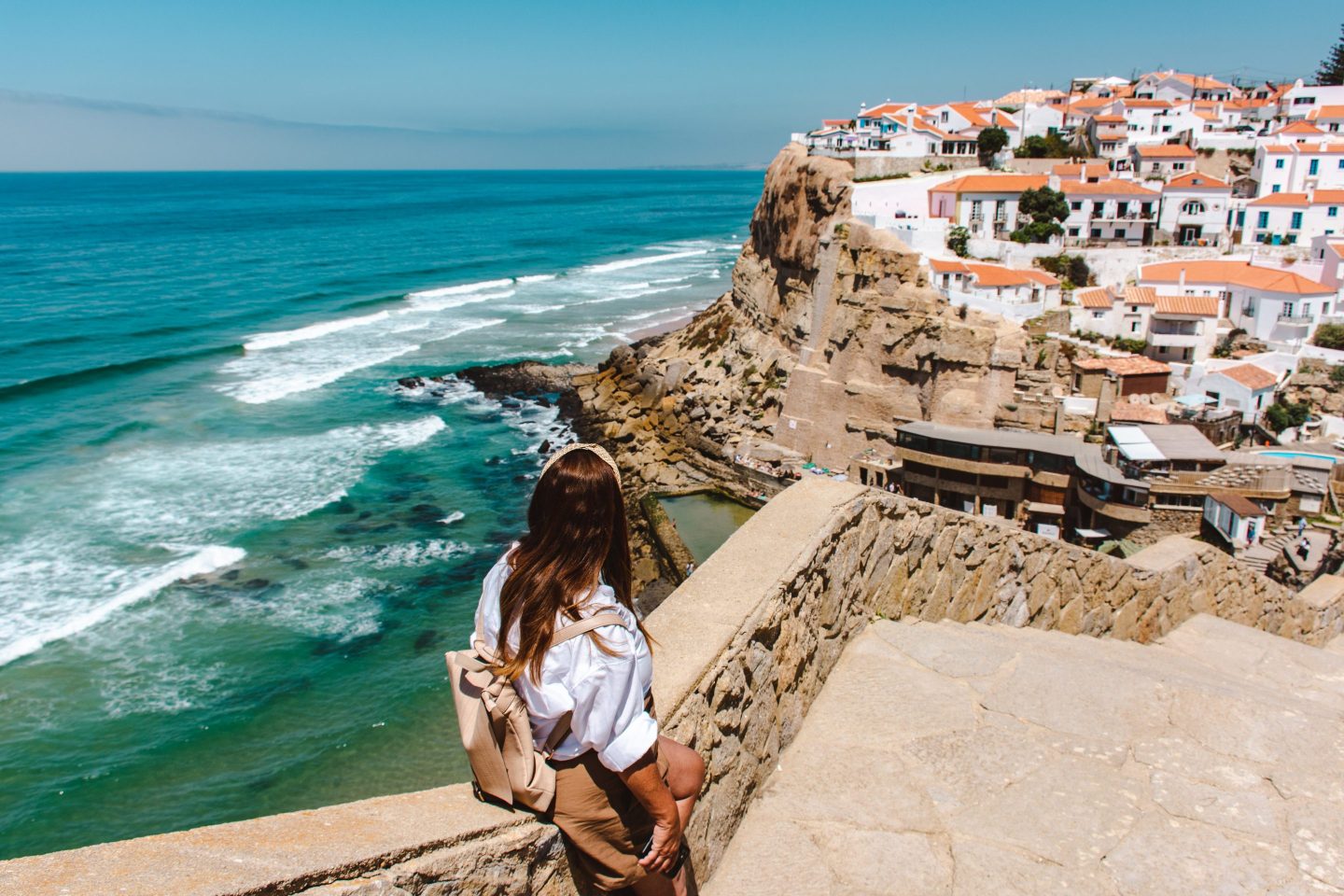 Rear view of woman sitting staring the view of the ocean and Azenhas do Mar town in Sintra, Lisbon, Portugal